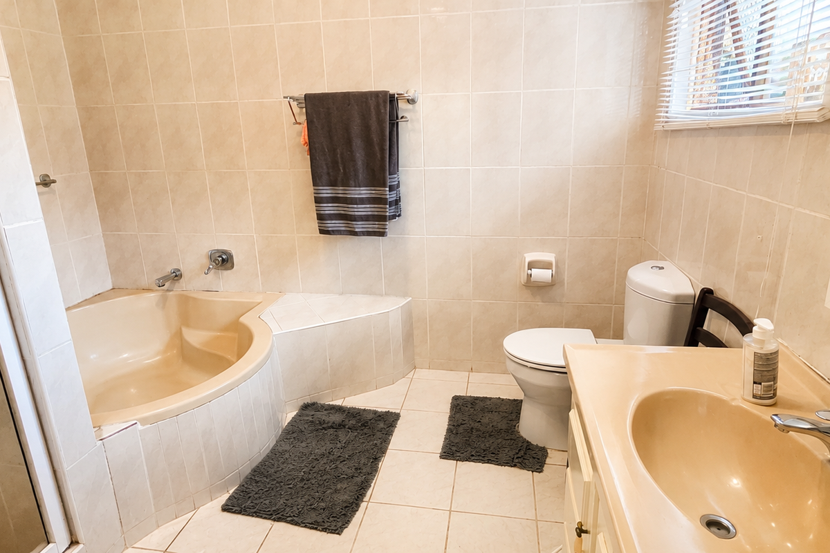 A functional bathroom featuring a built-in corner bathtub, vanity with integrated basin, toilet, and neutral wall and floor tiling.