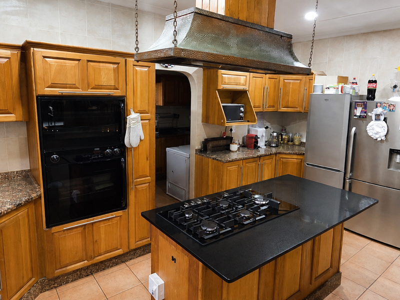 A spacious kitchen featuring warm wooden cabinetry, granite countertops, a central island with a gas hob, and a distinctive copper extractor hood.