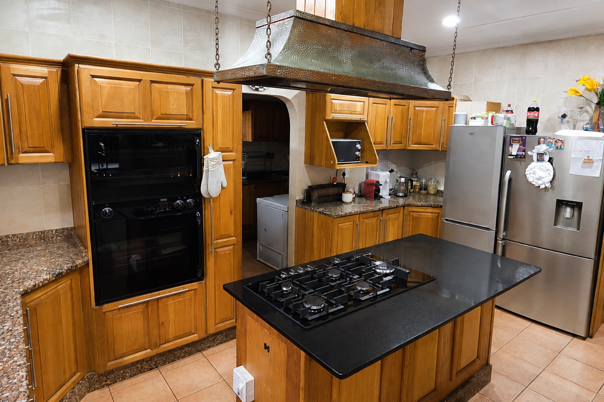 A spacious kitchen featuring warm wooden cabinetry, granite countertops, a central island with a gas hob, and a distinctive copper extractor hood.