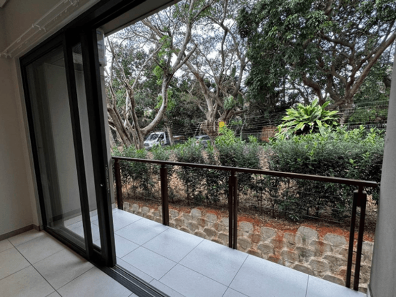 A clean, tiled balcony featuring a modern metal railing, accessible via large sliding glass doors, overlooking a landscaped garden area.