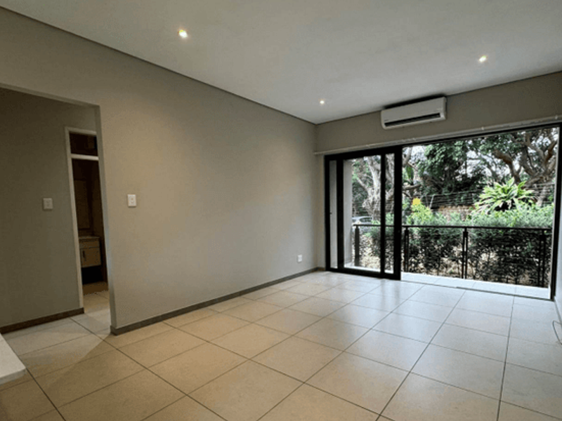 A bright, empty living room featuring neutral tiled floors, recessed lighting, a wall-mounted air conditioner, and large sliding doors leading to a private balcony.