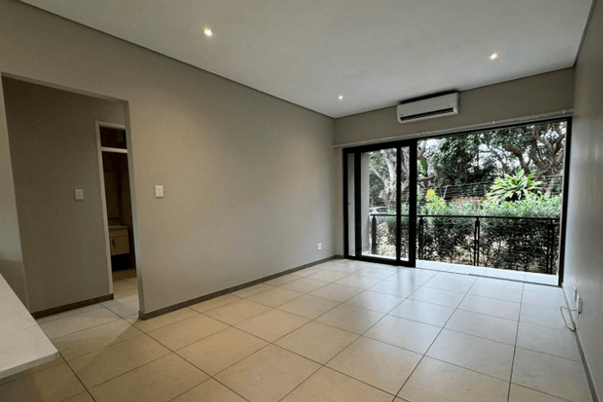A bright, empty living room featuring neutral tiled floors, recessed lighting, a wall-mounted air conditioner, and large sliding doors leading to a private balcony.