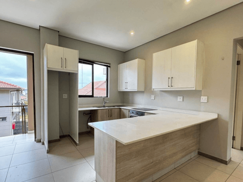 Modern kitchen featuring light wood-tone cabinetry, white stone countertops, integrated electric hob and oven, and tiled flooring with direct access to a balcony.