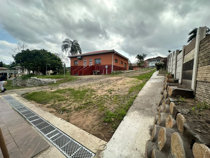 Exterior view of the property featuring a large unlandscaped yard, concrete retaining walls, drainage channels, and a tiled patio area leading to the main entrance.