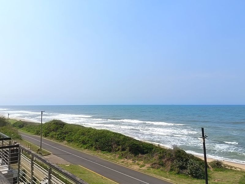 Elevated coastal view overlooking the ocean, beach, and adjacent coastal road from a balcony.
