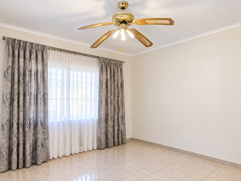 A bright, empty bedroom featuring neutral tiled flooring, a ceiling fan, and large windows with installed security bars.