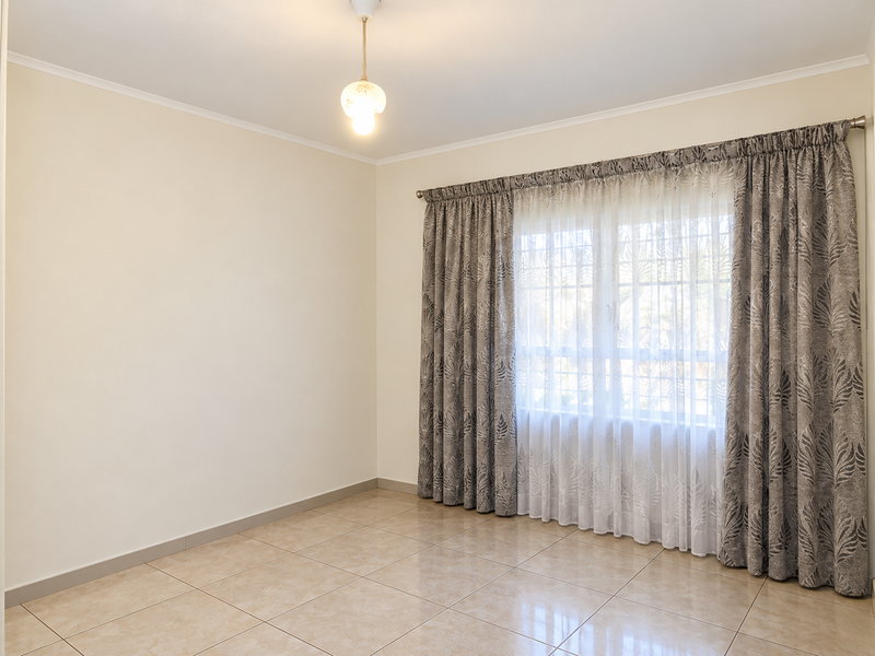 A bright and spacious empty bedroom featuring neutral tiled flooring, a central ceiling light, and a window dressed with decorative curtains.