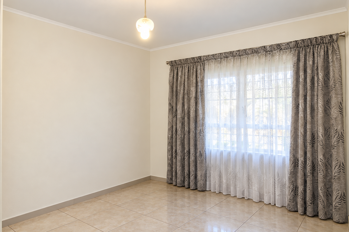 A bright and spacious empty bedroom featuring neutral tiled flooring, a central ceiling light, and a window dressed with decorative curtains.