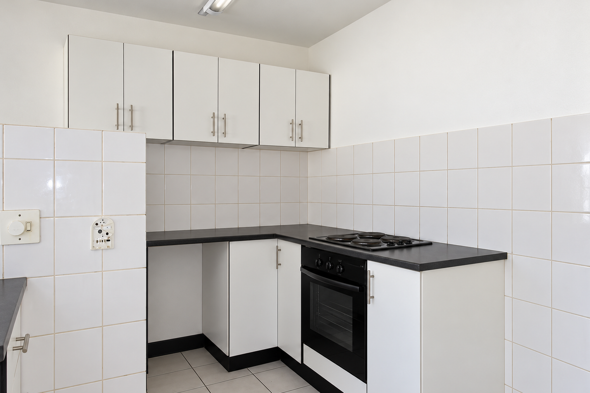 A functional, compact kitchen featuring white cabinetry, black countertops, a built-in oven and hob, and clean white tiled splashbacks and flooring.