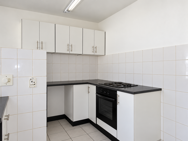 A functional, compact kitchen featuring white cabinetry, black countertops, a built-in oven and hob, and clean white tiled splashbacks and flooring.