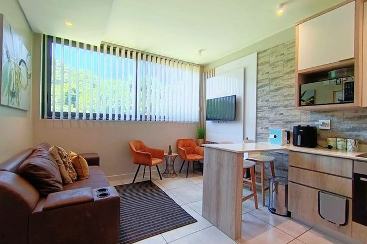 Modern open-plan living area featuring a compact kitchen with built-in appliances, a stone-cladding feature wall, and large windows with vertical blinds.