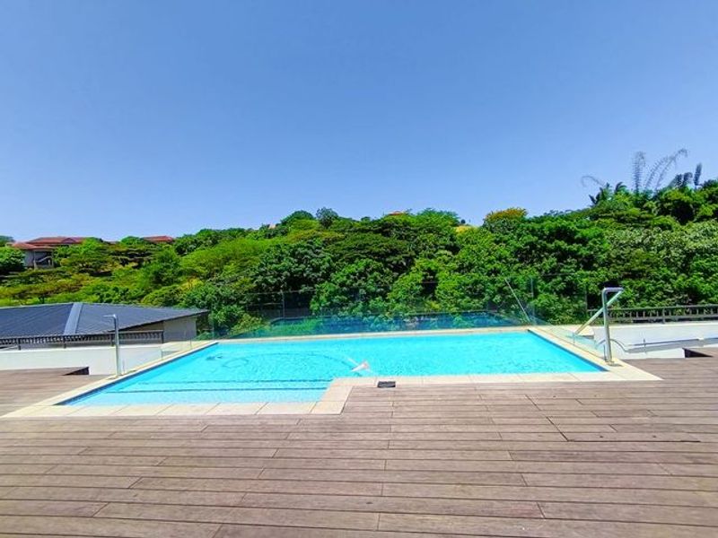 A well-maintained outdoor swimming pool area featuring timber decking, a glass balustrade, and scenic hillside views.