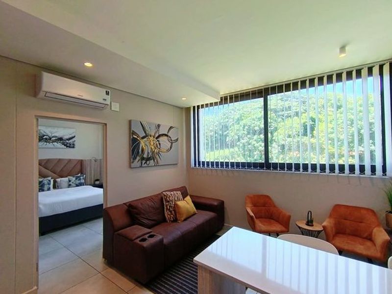 A bright open-plan living area featuring tiled floors, recessed lighting, a split-system air conditioner, and large windows with vertical blinds overlooking greenery.