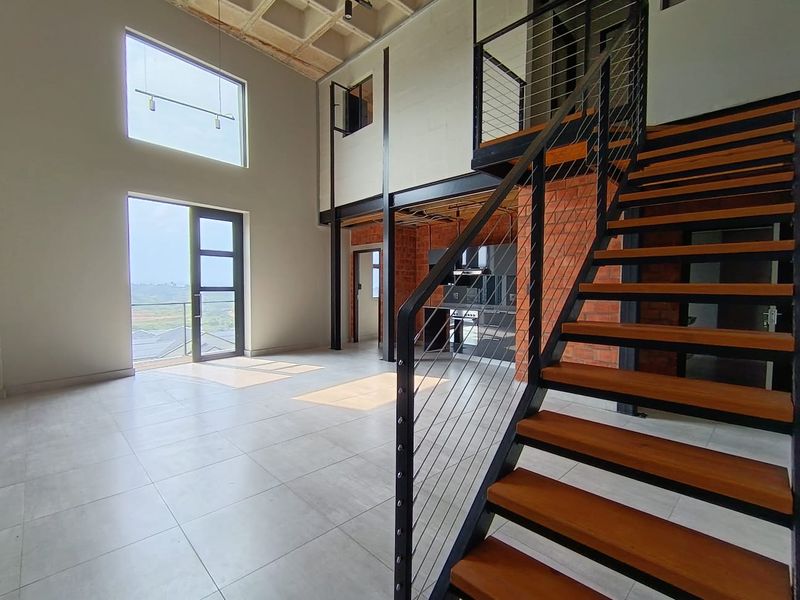 Spacious open-plan living area featuring high ceilings, exposed brickwork, a modern steel and timber staircase, and large windows providing ample natural light.