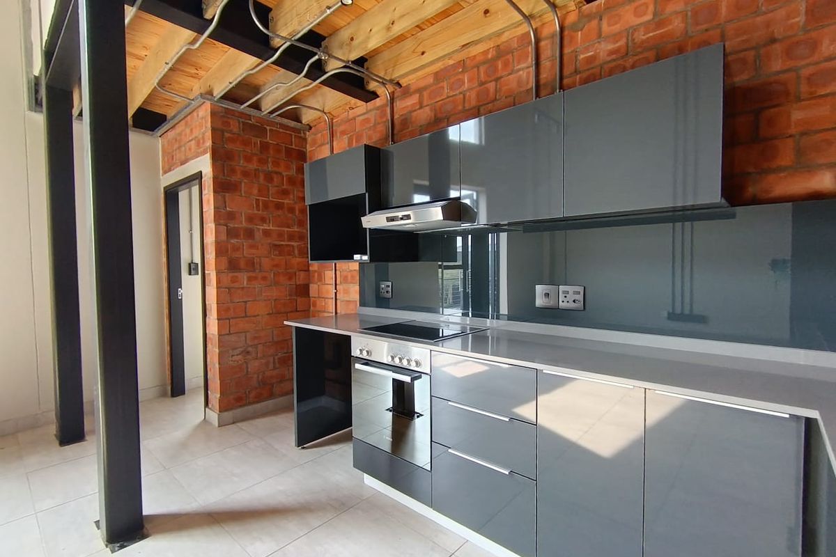 Modern industrial-style kitchen featuring exposed brick walls, timber ceiling beams, high-gloss grey cabinetry, and integrated appliances with a sleek stone countertop.