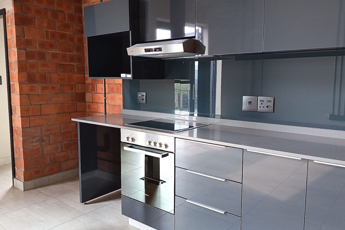 Modern kitchen featuring sleek grey cabinetry, integrated oven and hob, glass splashback, and a striking exposed brick feature wall.
