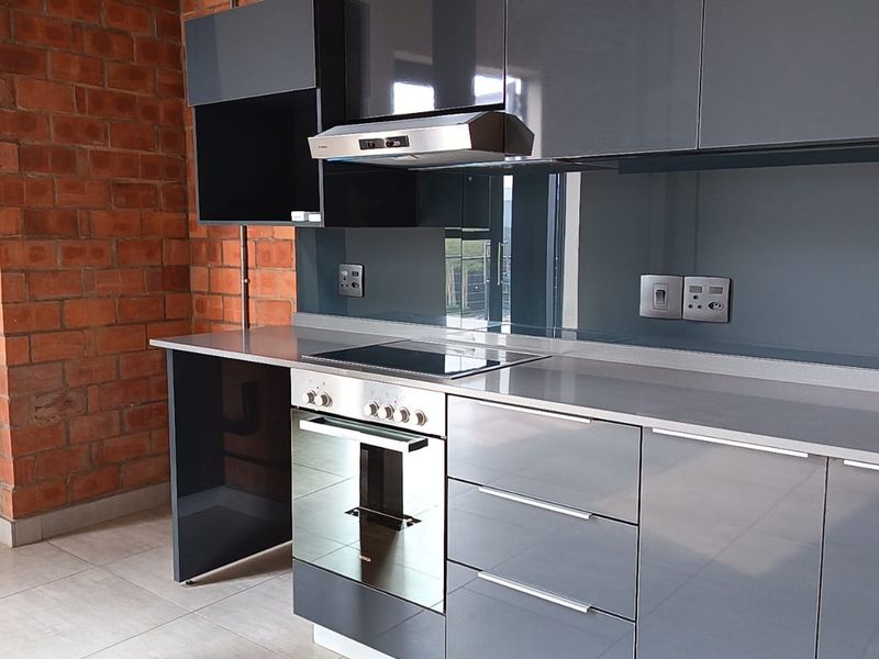 Modern kitchen featuring sleek grey cabinetry, integrated oven and hob, glass splashback, and a striking exposed brick feature wall.