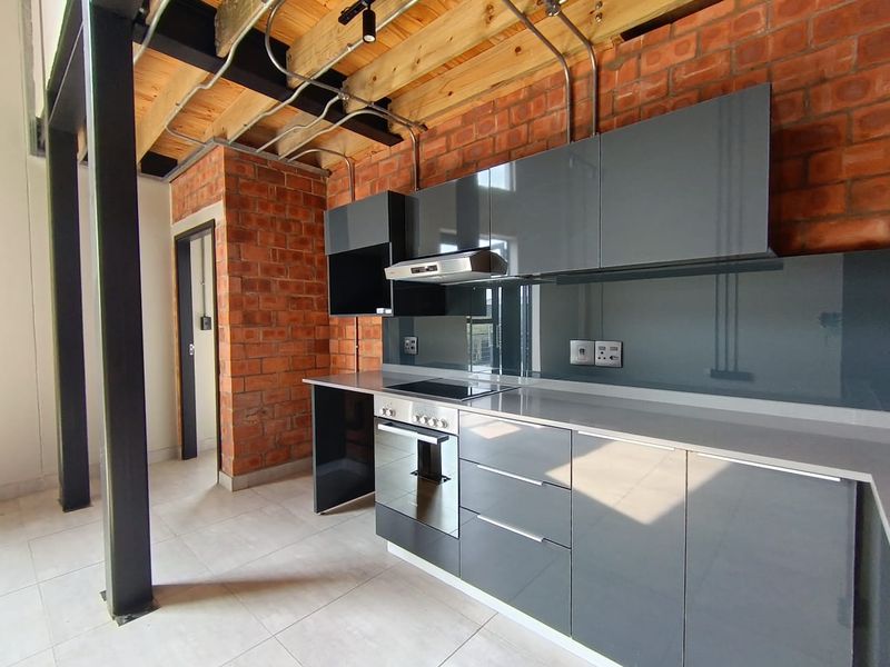 Modern industrial-style kitchen featuring exposed brick walls, timber ceiling beams, high-gloss grey cabinetry, and integrated appliances with a sleek stone countertop.
