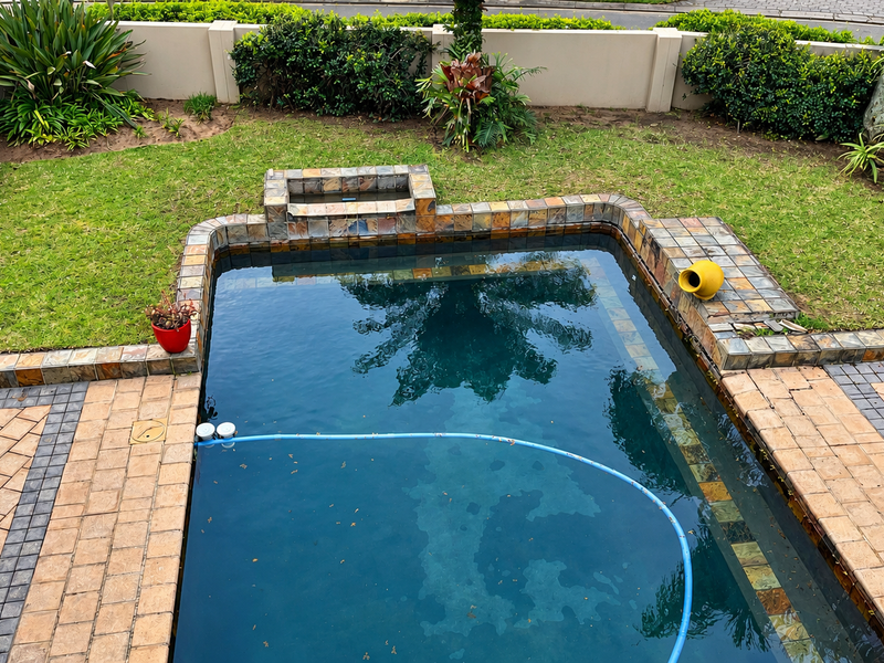 A well-maintained swimming pool area featuring stone coping, paved surround, and a landscaped garden enclosed by a boundary wall.