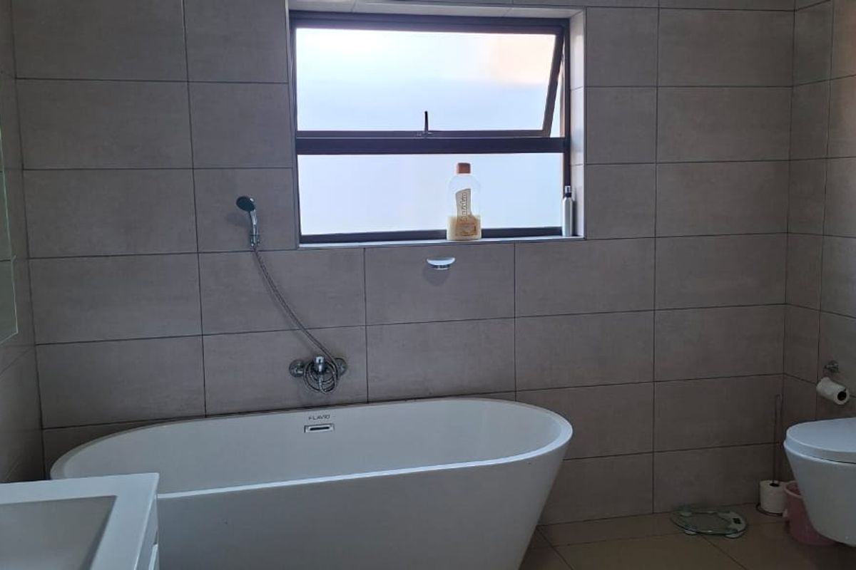 Modern bathroom featuring a freestanding bathtub, wall-mounted toilet, vanity basin, and neutral floor-to-ceiling tiling with a top-hung window for natural light.