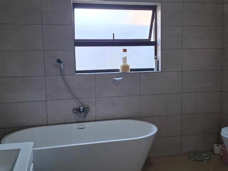 Modern bathroom featuring a freestanding bathtub, wall-mounted toilet, vanity basin, and neutral floor-to-ceiling tiling with a top-hung window for natural light.