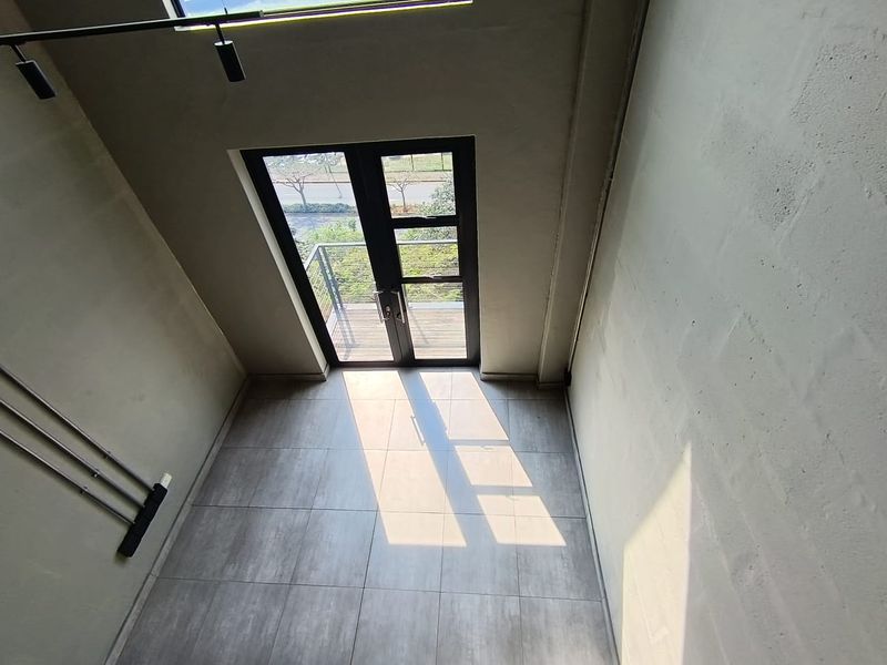 A bright, double-volume living area featuring modern tiled floors, exposed blockwork walls, and double glass doors leading to an outdoor balcony.