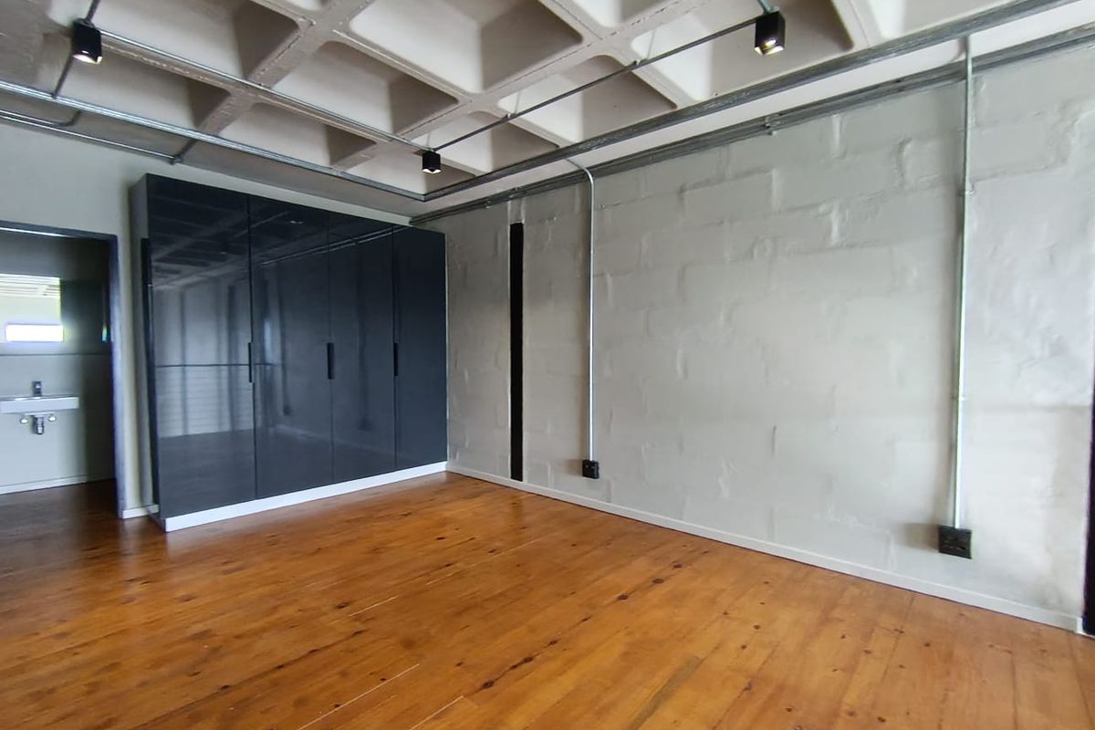 Spacious bedroom featuring industrial-style exposed concrete waffle ceiling, timber flooring, built-in cupboards, and exposed electrical conduit with track lighting.