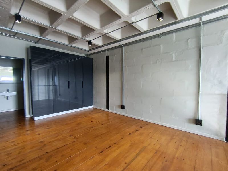Spacious bedroom featuring industrial-style exposed concrete waffle ceiling, timber flooring, built-in cupboards, and exposed electrical conduit with track lighting.