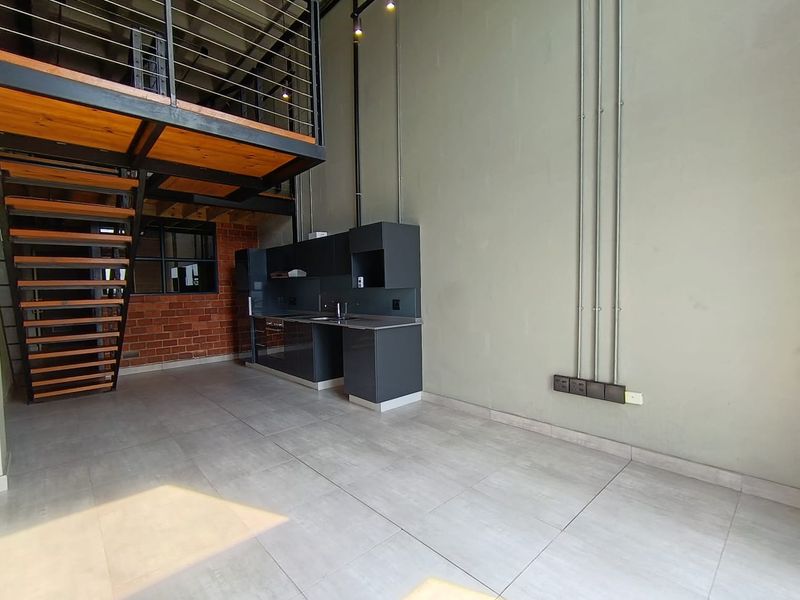Modern industrial-style open plan living area featuring a mezzanine level, exposed brickwork, sleek kitchen cabinetry, and tiled flooring in a spacious double-volume layout.