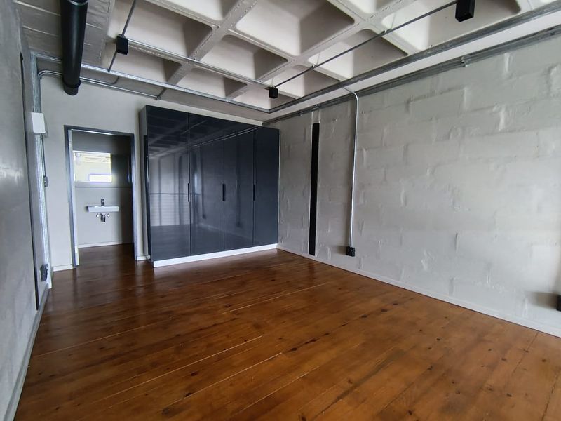 Modern industrial-style bedroom featuring exposed concrete waffle ceiling, timber flooring, painted brick walls, and a sleek built-in wardrobe with an adjoining en-suite bathroom.
