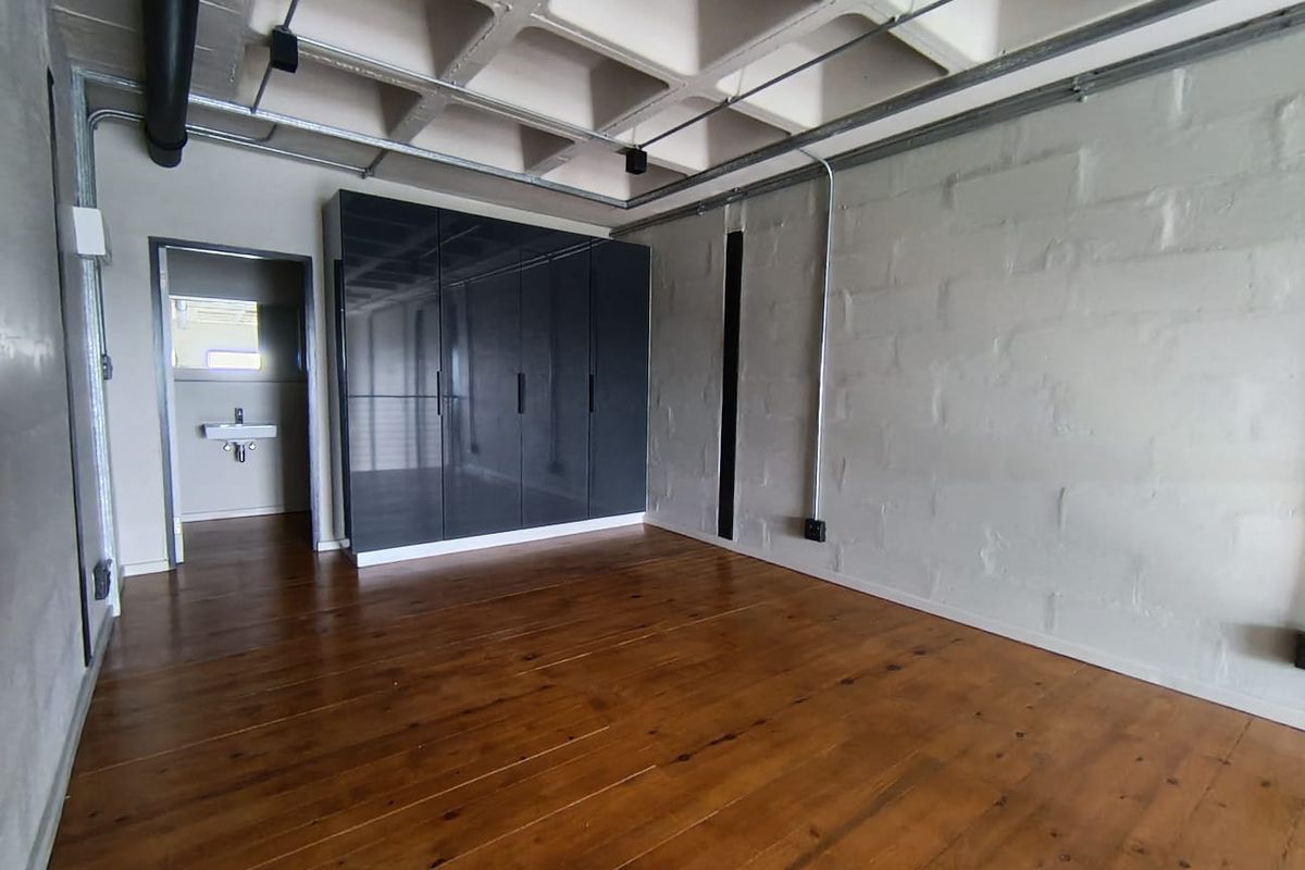 Modern industrial-style bedroom featuring exposed concrete waffle ceiling, timber flooring, painted brick walls, and a sleek built-in wardrobe with an adjoining en-suite bathroom.