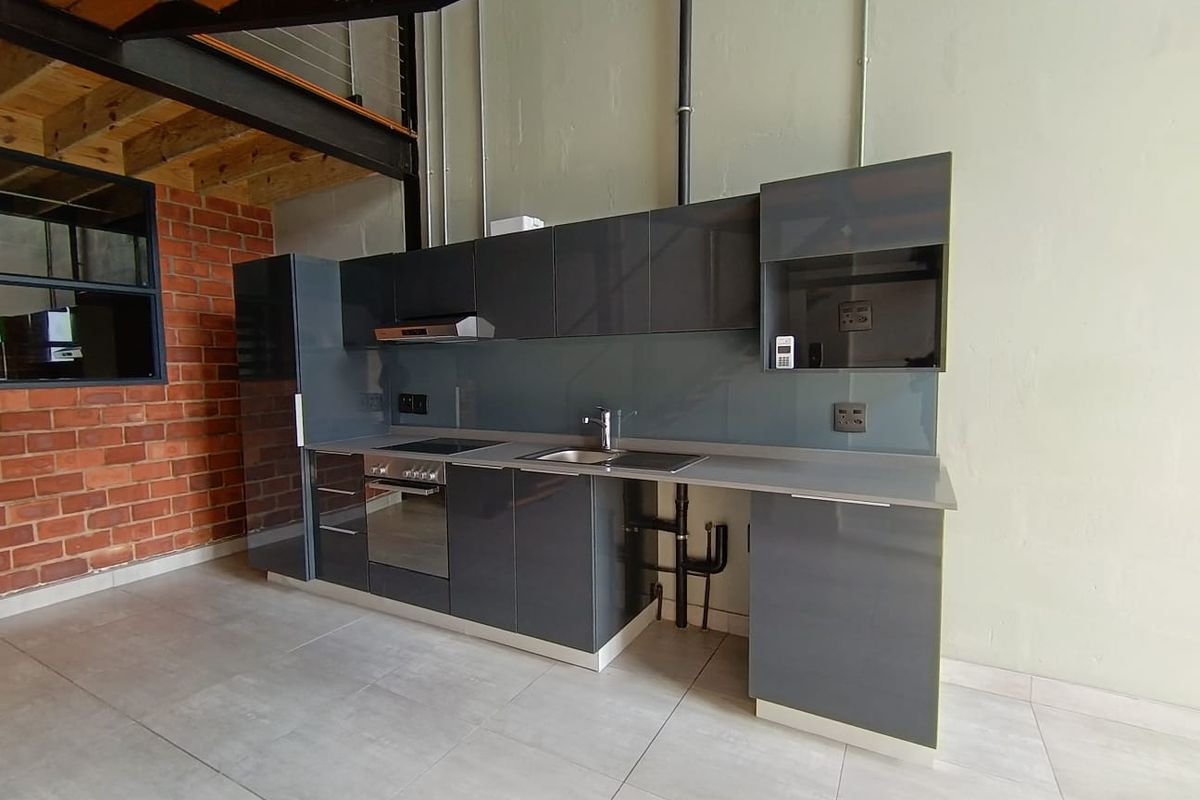 Modern industrial-style kitchen featuring sleek grey cabinetry, integrated appliances, exposed brickwork, and steel structural elements in an open-plan apartment setting.