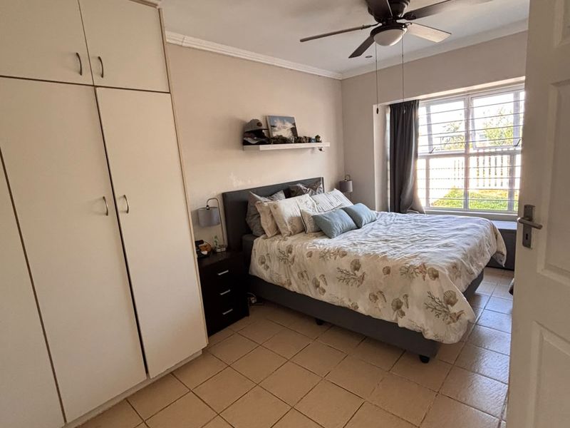 A bedroom featuring built-in cupboards, tiled flooring, and a ceiling fan, with security bars installed on the window.