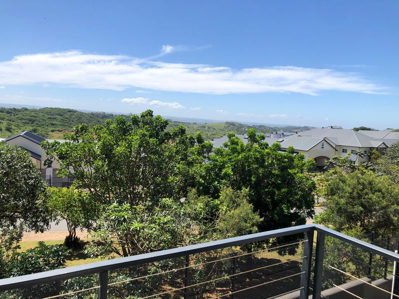 Elevated view from a balcony overlooking the surrounding residential estate, lush greenery, and distant hills under a clear blue sky.