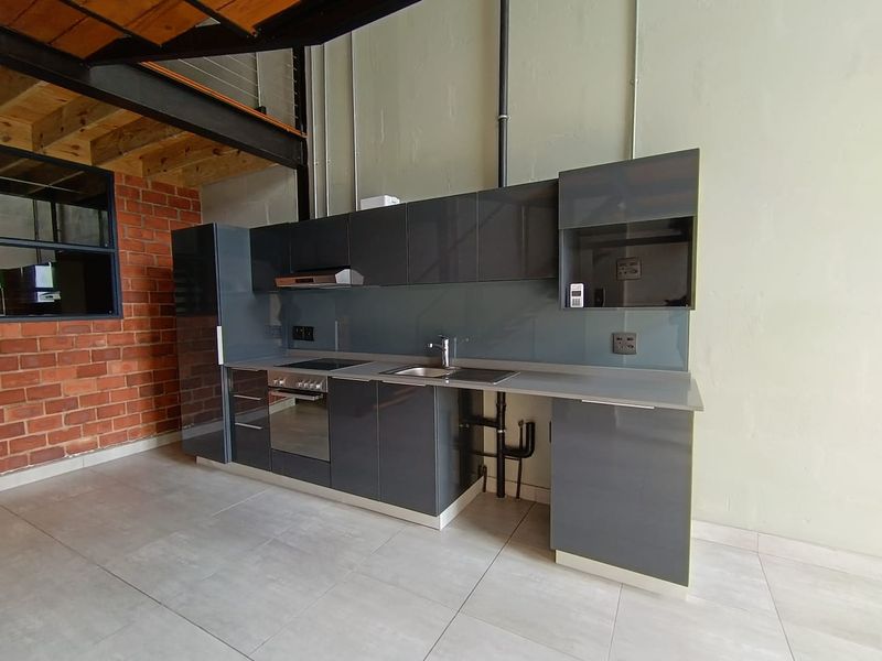 Modern industrial-style kitchen featuring sleek grey cabinetry, integrated appliances, exposed brickwork, and steel structural elements in an open-plan apartment setting.