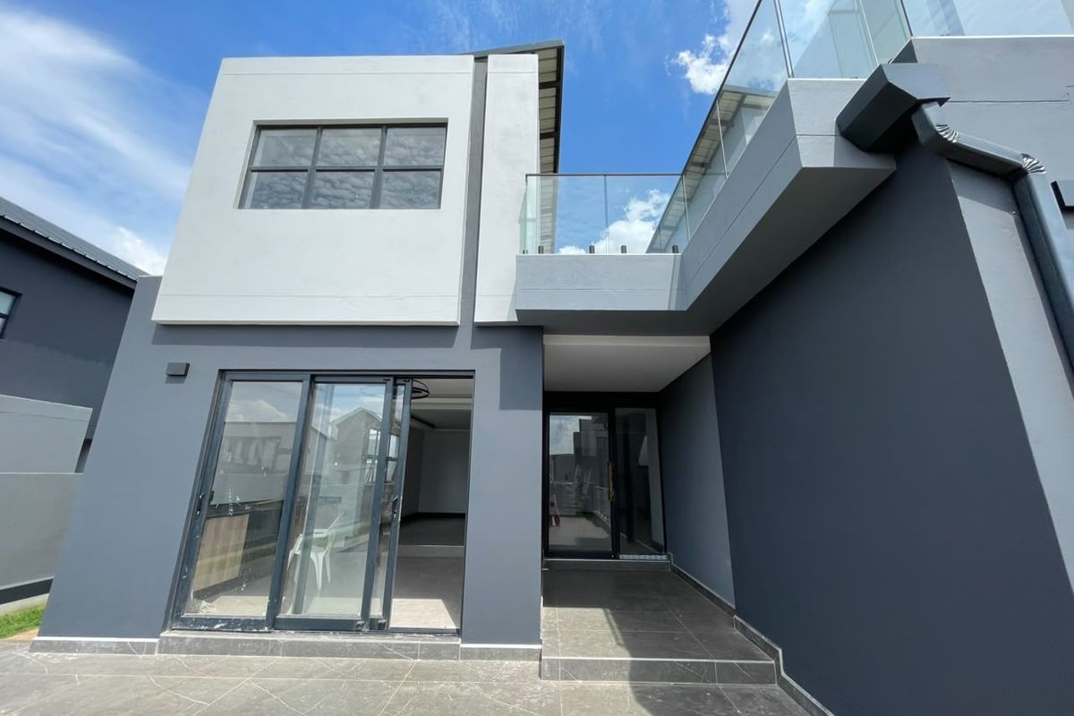 Modern rear exterior view of the house featuring a tiled patio, glass balustrade balcony, and dark grey rendered facade with large sliding glass doors.