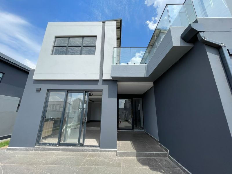 Modern rear exterior view of the house featuring a tiled patio, glass balustrade balcony, and dark grey rendered facade with large sliding glass doors.