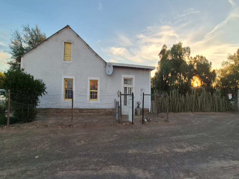 Front view of a single-story house featuring a stucco facade, corrugated metal roof, and perimeter wire fencing in a rural setting.