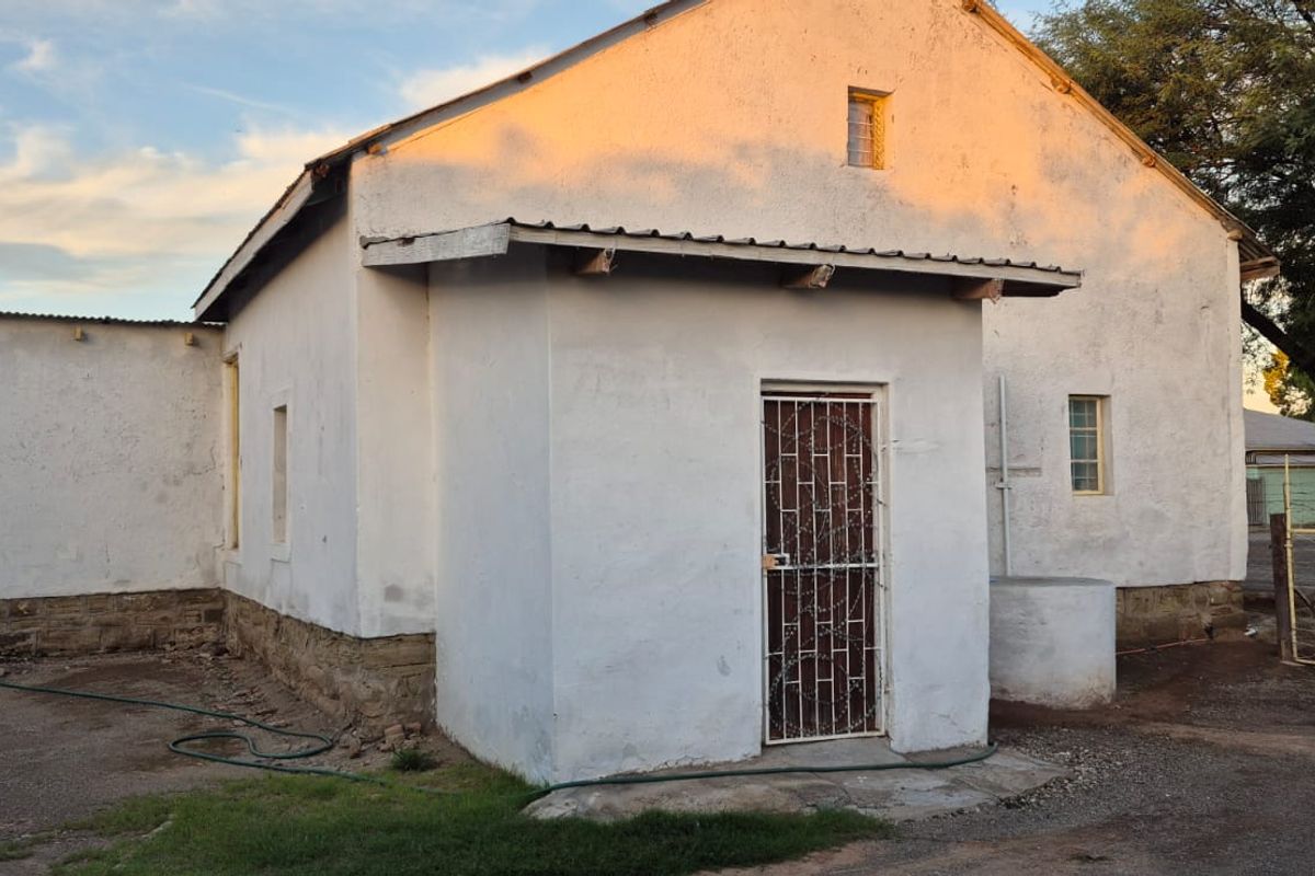 Rear view of a single-story house featuring plastered masonry walls, a corrugated metal roof, and a security gate entrance.