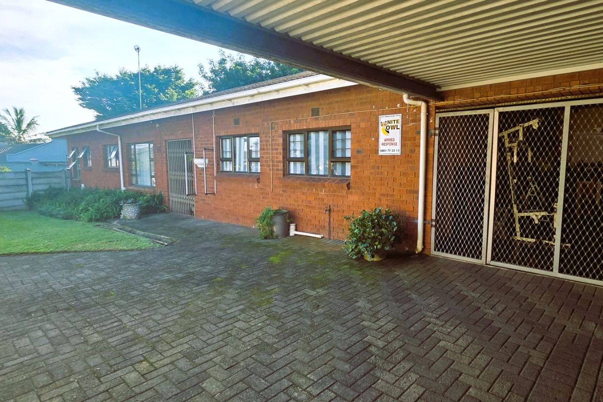Rear exterior view featuring a brick facade, paved driveway, covered carport area, and a small lawn with perimeter walling.