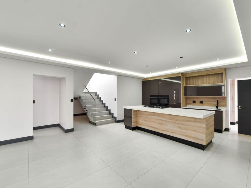 Modern open-plan kitchen featuring a central island, built-in appliances, sleek cabinetry, and a contemporary staircase with glass balustrade in a bright, minimalist space.