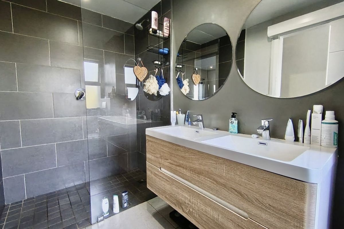 Modern bathroom featuring a double vanity with wood-finish cabinetry, large circular mirrors, and a spacious walk-in shower with dark grey tiling.