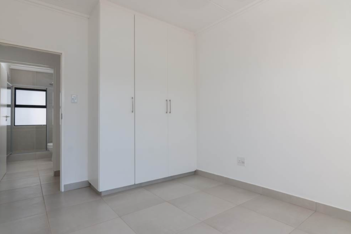 A clean, empty bedroom featuring neutral tiled flooring and white built-in cupboards, with an adjacent view into a tiled bathroom.