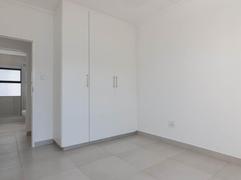 A clean, empty bedroom featuring neutral tiled flooring and white built-in cupboards, with an adjacent view into a tiled bathroom.