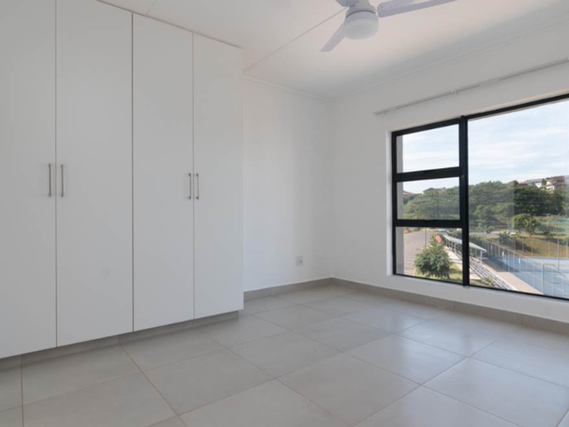 A bright, empty bedroom featuring neutral tiled flooring, built-in white cupboards, a ceiling fan, and a large window with views of the surrounding area.