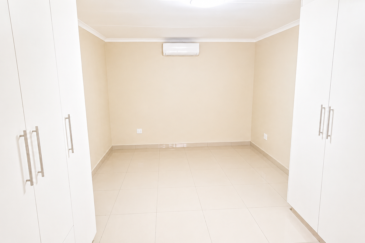 A clean, empty bedroom featuring built-in white wardrobes, tiled flooring, and a wall-mounted air conditioning unit.