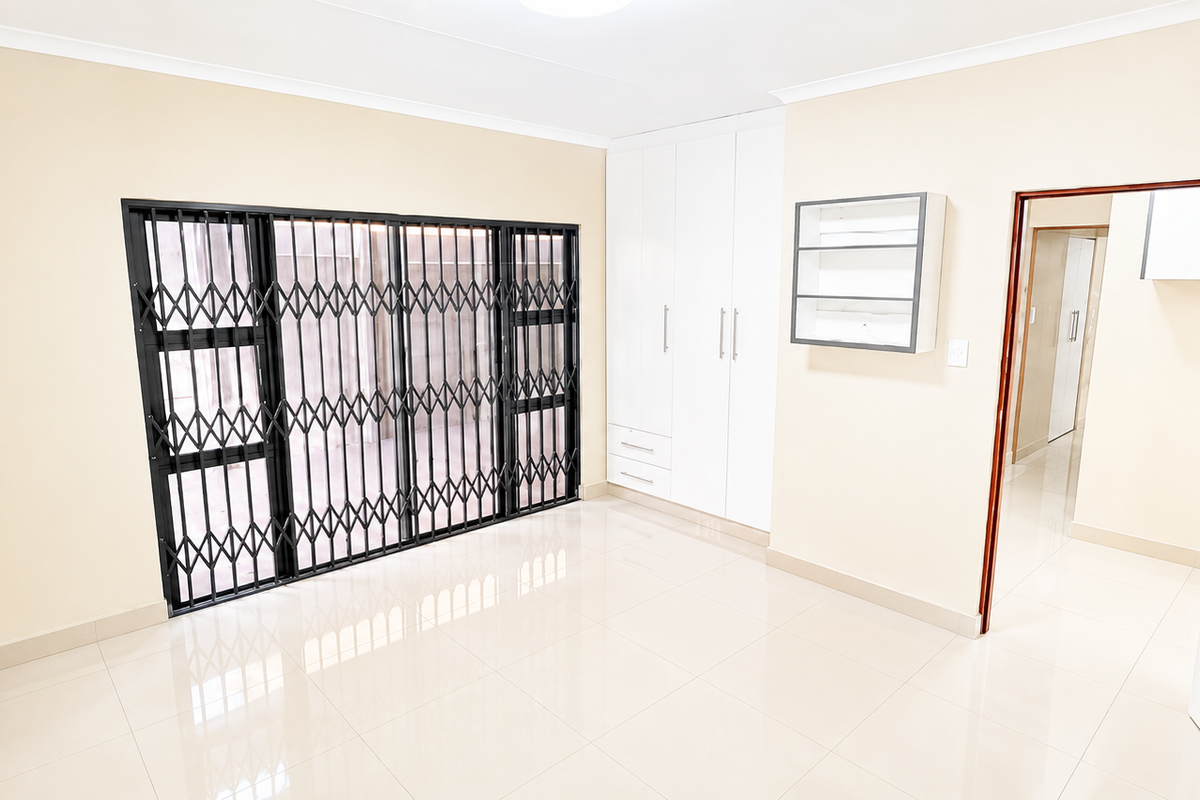 A bright, empty bedroom featuring tiled flooring, built-in white cabinetry, and a secure metal sliding gate over the glass door.