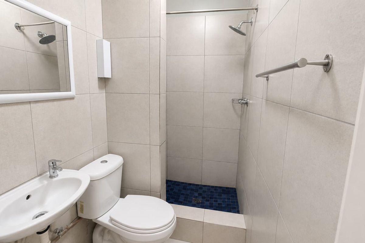 A clean, functional bathroom featuring neutral wall tiles, a walk-in shower with blue mosaic floor tiling, a wall-mounted basin, and a standard toilet.