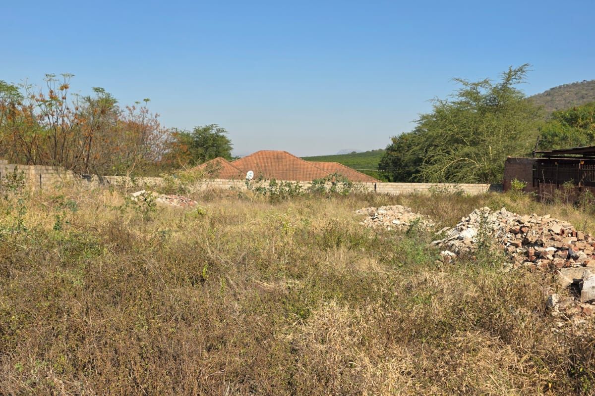 A vacant residential plot featuring overgrown vegetation and scattered rubble, enclosed by a perimeter wall with surrounding trees and clear sky views.