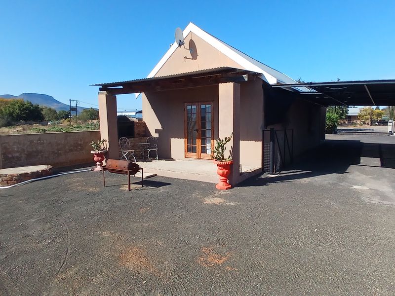 Front view of a single-story house featuring a covered patio with a built-in braai, attached carport, and gravel driveway.
